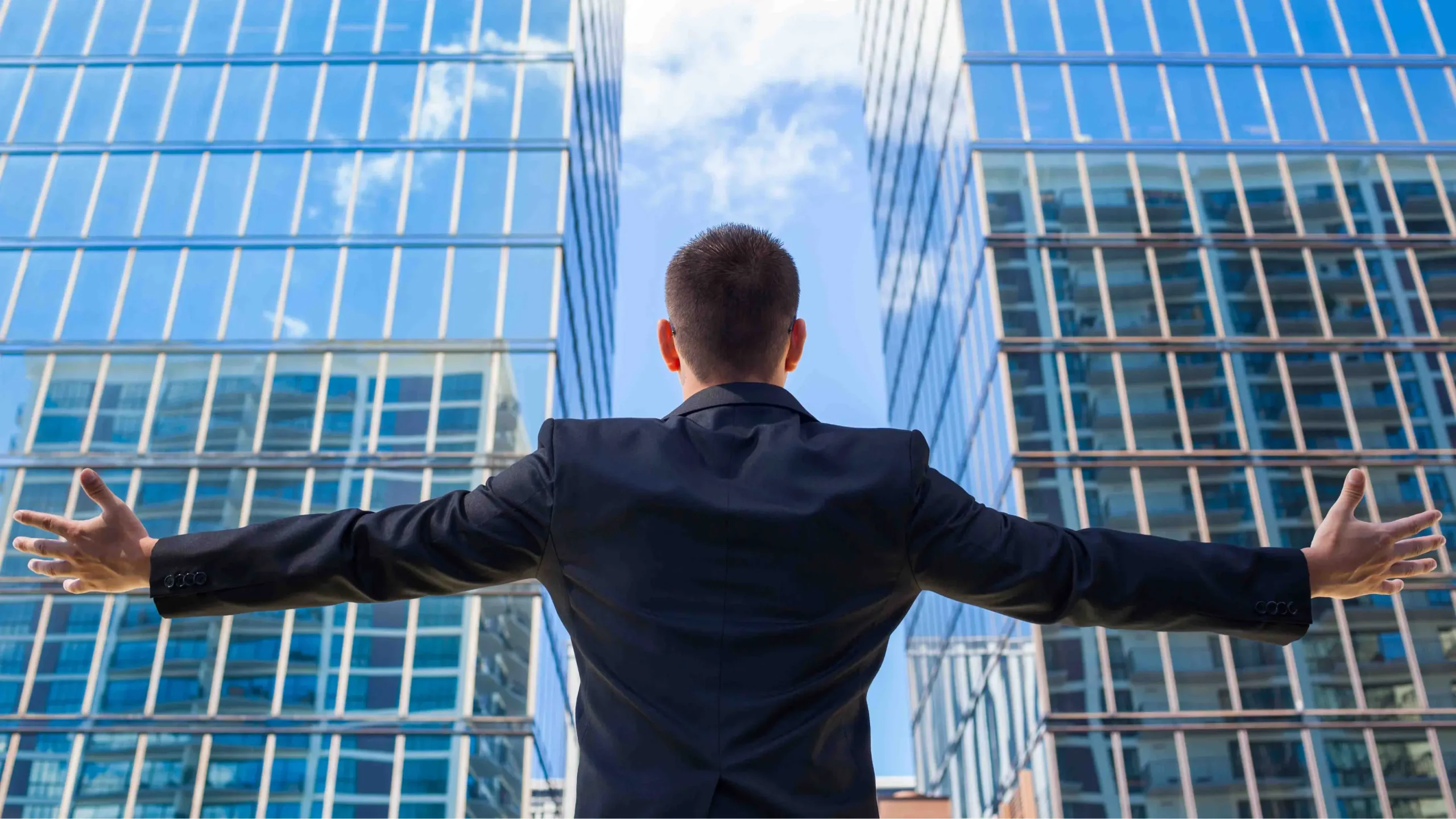 Person in a suit with open arms looking at the sky between two buildings.