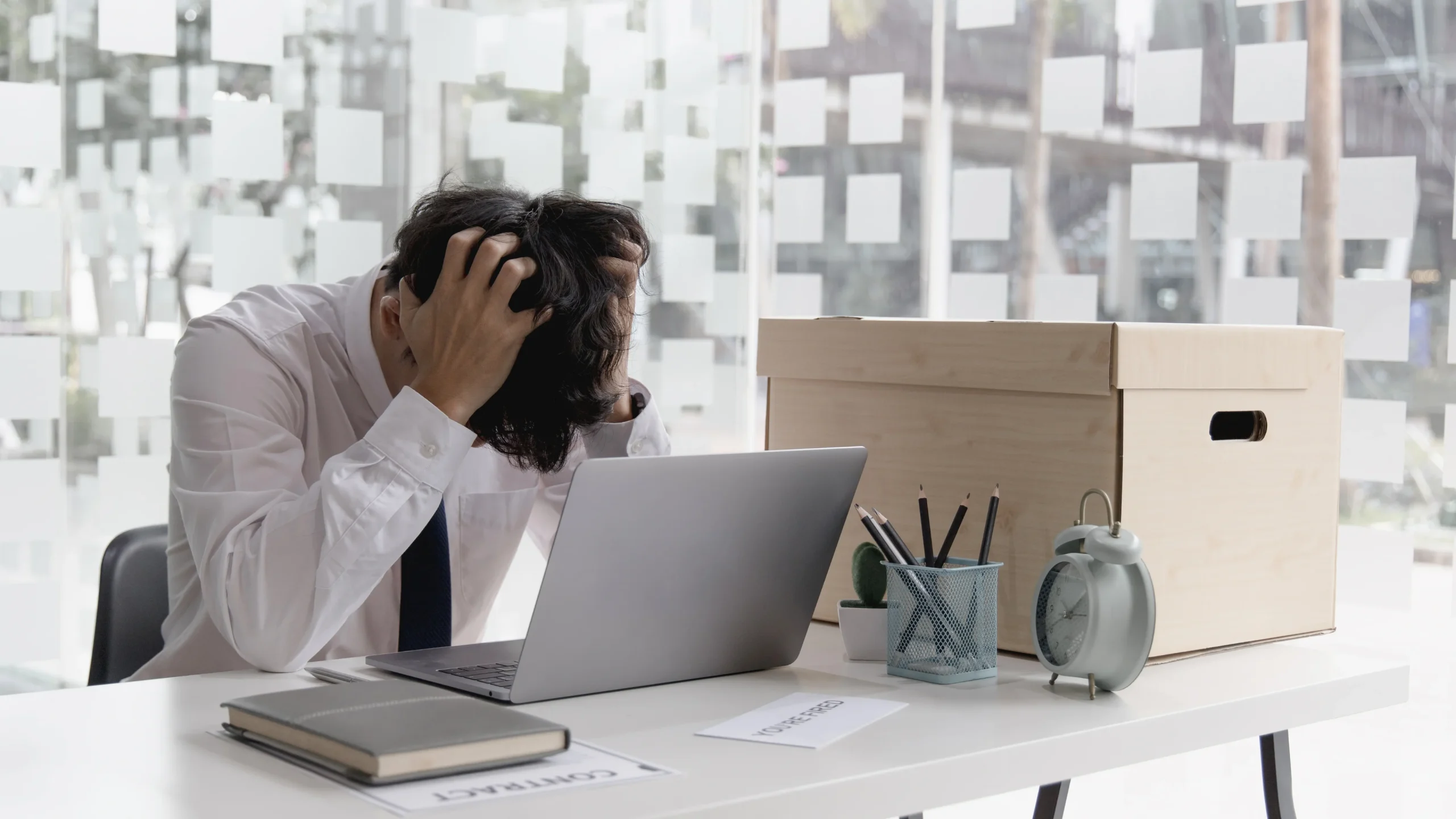 Frustrated businessman sitting at a desk with his head in his hands, surrounded by office supplies, a laptop, and a contract document, symbolizing stress or job loss.