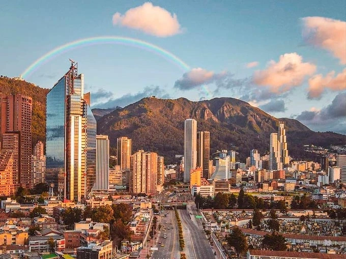 A view of Bogotá with modern skyscrapers, lush mountains in the background, and a rainbow arching over the city.