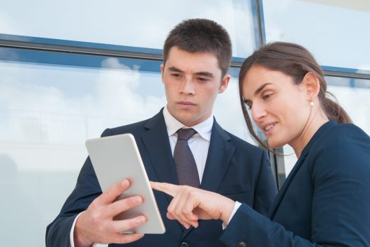 Focused coworkers watching presentation outside office. Young man and woman in office suits standing against outdoor glass wall and using tablet. Interaction concept