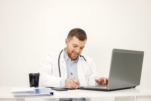 Male doctor with laptop at desk in medical office. Man wearing white medical gown. Takeaway coffee standing on a table.