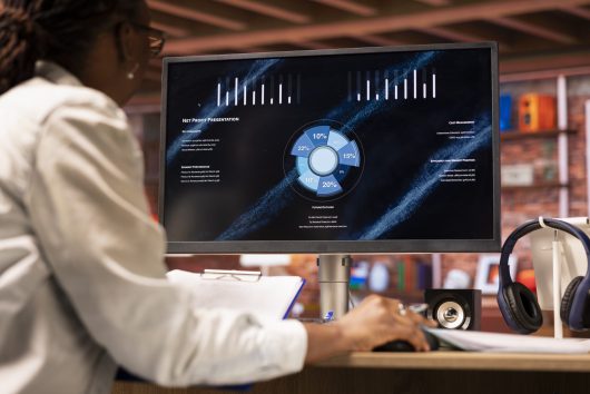 Woman watching business conference on computer screen, looking over statistics on portfolio documents. African american person in apartment looking at business presentation